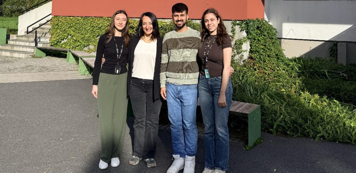 Üç genç mezun (iki kadın, bir erkek) ön planda gülümseyerek poz veriyor. Arka planda Norveç’te yeşil doğa manzarası ve modern üniversite binası yer almakta. Staj için buradalar.